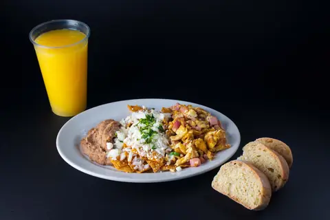 Plato de chilaquiles rojos con queso, huevo revuelto con jamón y frijoles, con pan y jugo de naranja